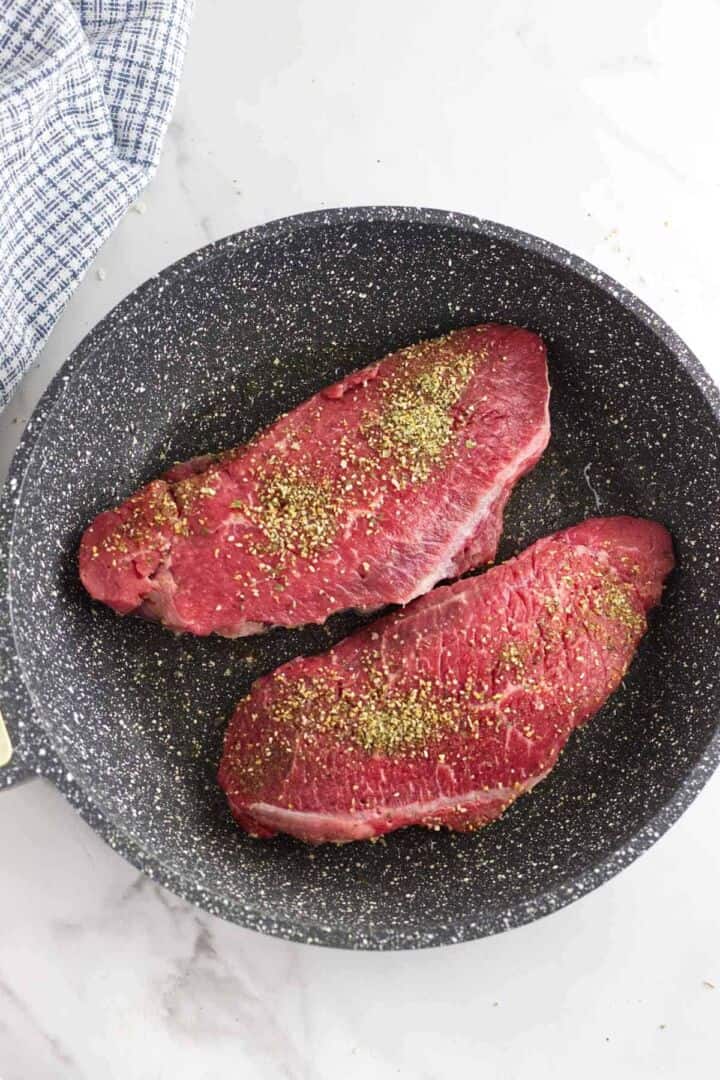 A nonstick frying pan with two raw, seasoned steak cuts inside, on a white marble surface. A blue and white checkered cloth is partially visible in the top left corner.