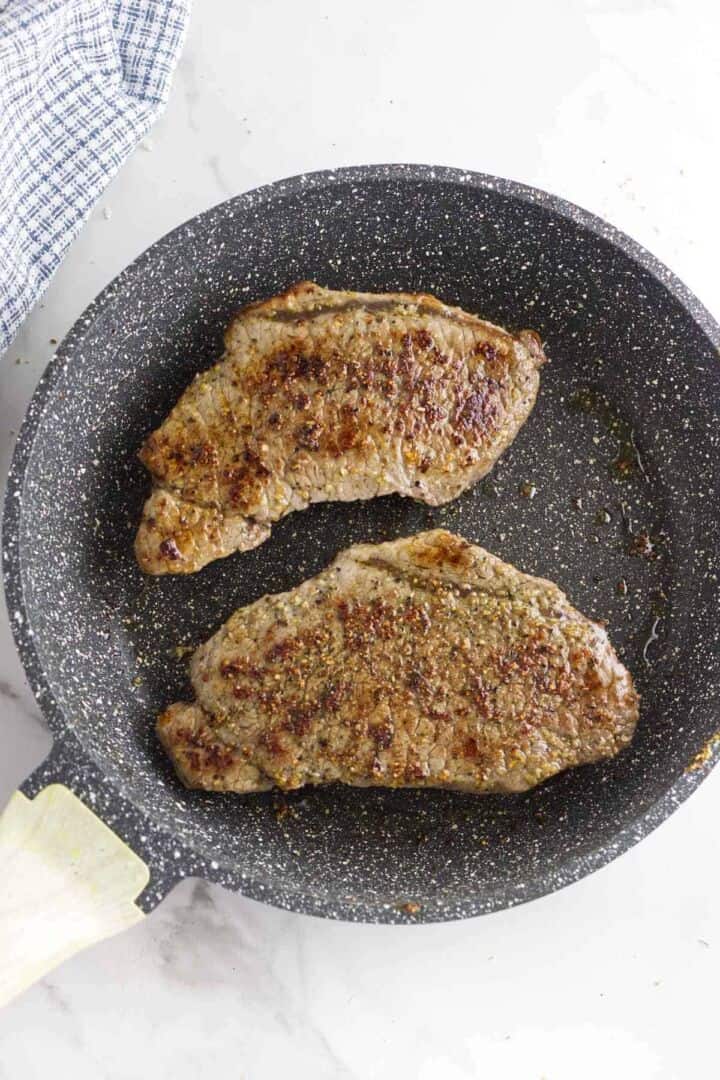 Two cooked steaks with a browned, seasoned crust are in a speckled black frying pan on a white surface. A blue and white checkered cloth is partially visible beside the pan.