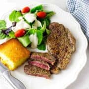 A plate with a grilled Denver steak, sliced to show a pink center, a green salad with grape tomatoes and creamy dressing, and a square piece of cornbread. A fork and knife are beside the plate.