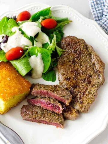 A plate with a grilled Denver steak, sliced to show a pink center, a green salad with grape tomatoes and creamy dressing, and a square piece of cornbread. A fork and knife are beside the plate.