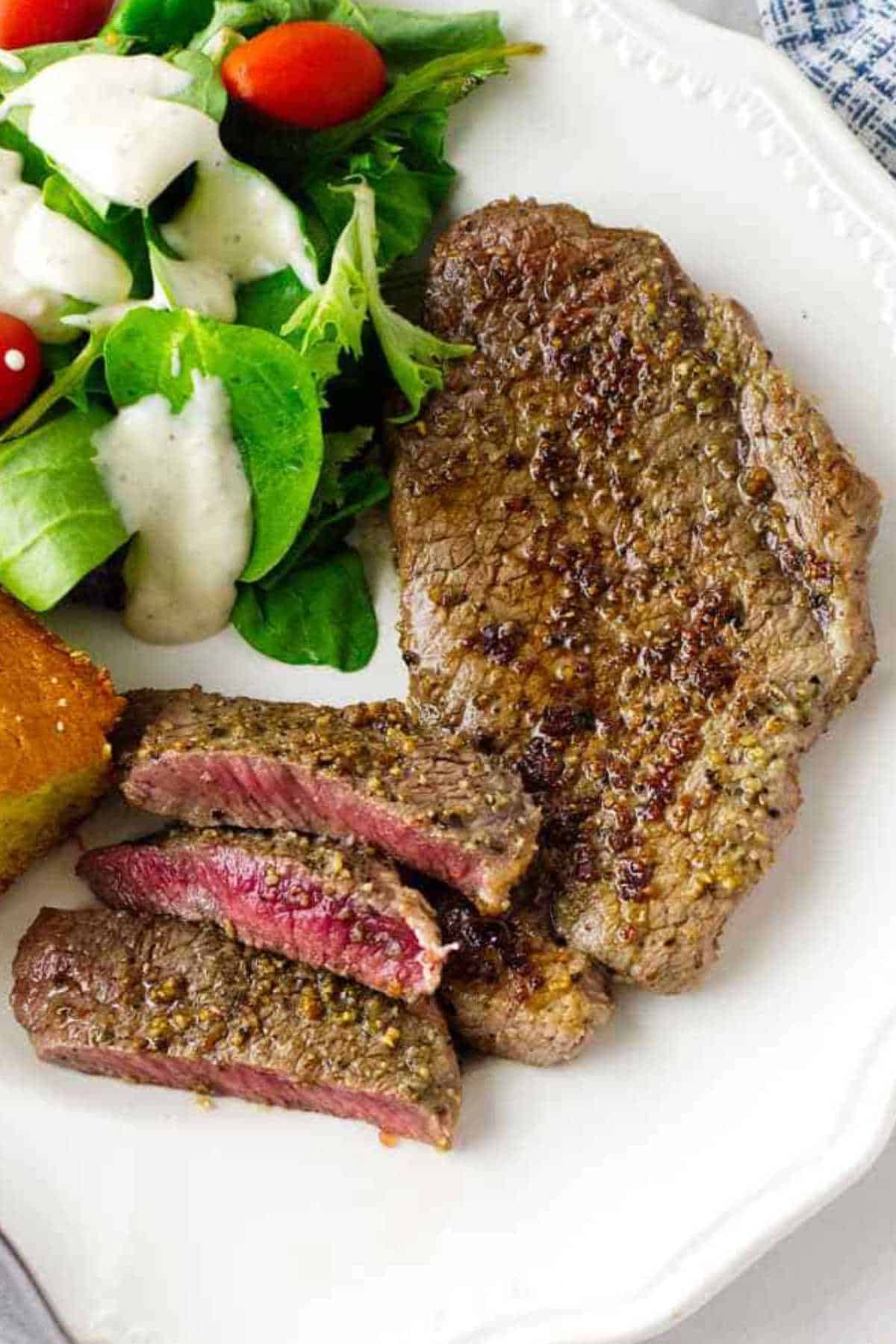 A plate with a grilled Denver steak, sliced to show a pink center, a green salad with grape tomatoes and creamy dressing, and a square piece of cornbread. A fork and knife are beside the plate.