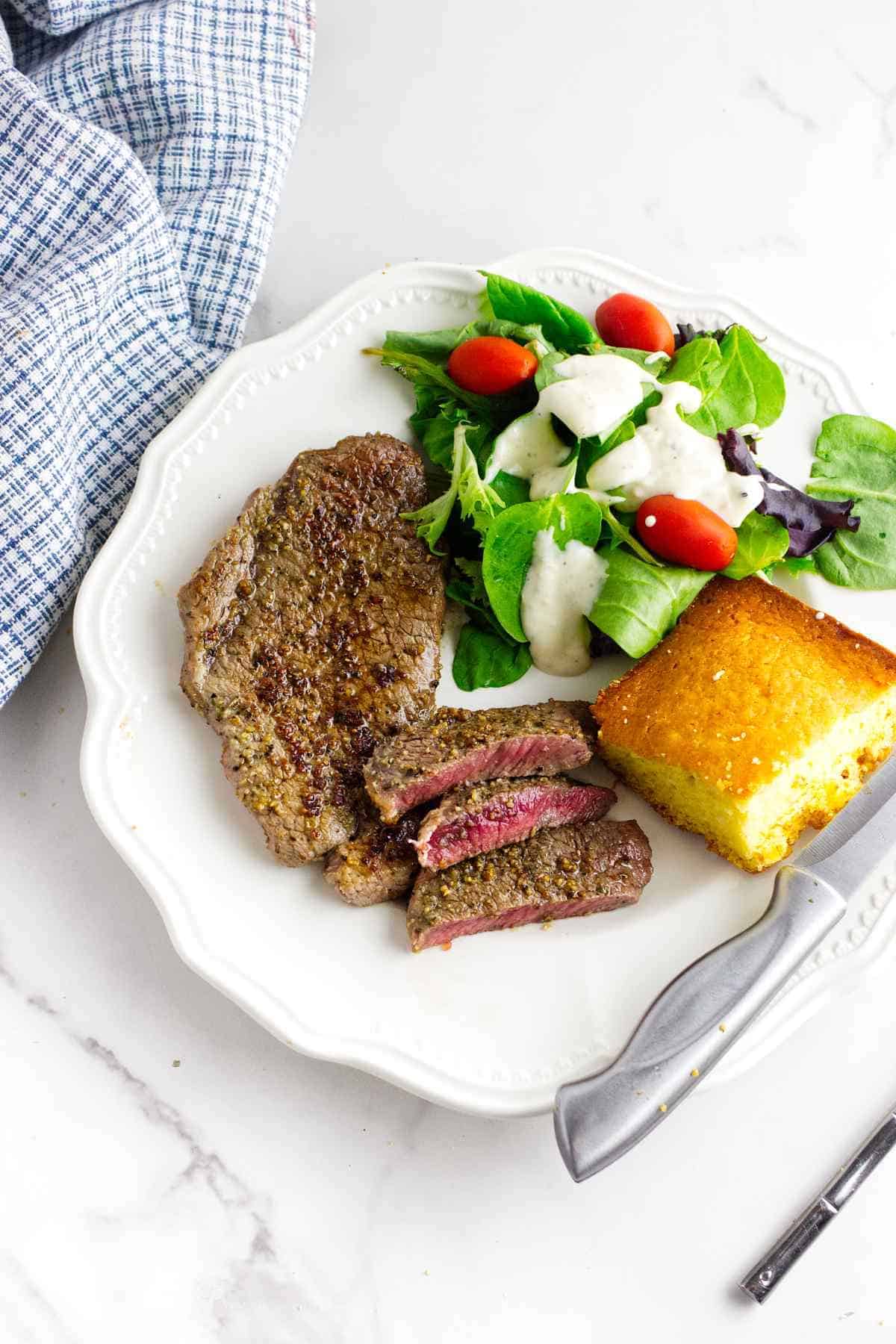 A white plate with a cooked steak, some slices cut, a square of cornbread, and a green salad with grape tomatoes and creamy dressing. A knife rests on the plate; a blue checked towel is nearby.