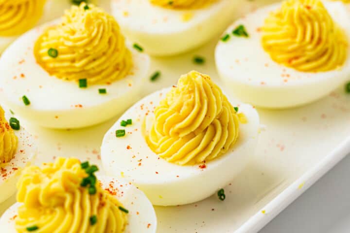 A close-up of devuled eggs on a white platter, with creamy yellow yolk filling piped into halved egg whites, garnished with chopped chives and a sprinkle of paprika.