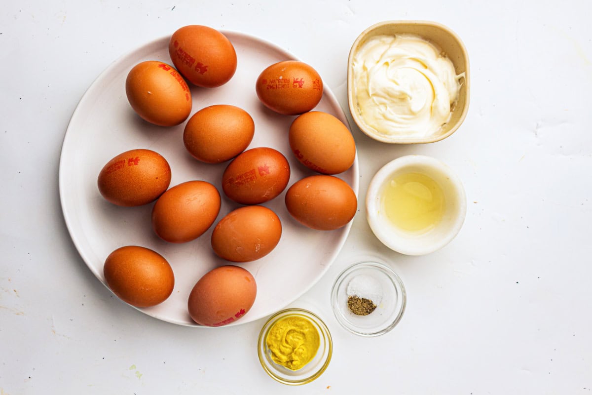 A plate with ten brown eggs sits next to small bowls of mayonnaise, vinegar, mustard, and a mix of salt and pepper—everything you need to make delicious devuled eggs—all arranged on a white surface.