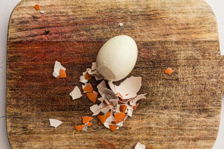 A peeled hard-boiled egg sits on a wooden cutting board next to pieces of broken brown eggshell, ready to be transformed into delicious deviled eggs.
