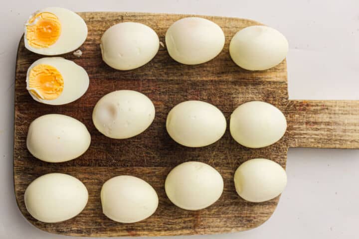 Eleven whole peeled hard-boiled eggs and one halved egg with a yellow yolk-perfect for making devuled eggs-are arranged on a wooden cutting board with a handle, set against a plain white background.