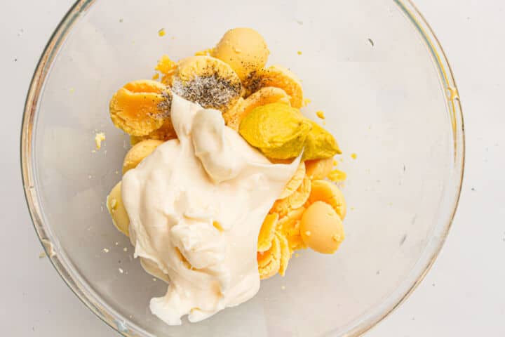 A glass bowl containing hard-boiled egg yolks, mayonnaise, yellow mustard, and black pepper, ready to be mixed for classic devuled eggs filling.