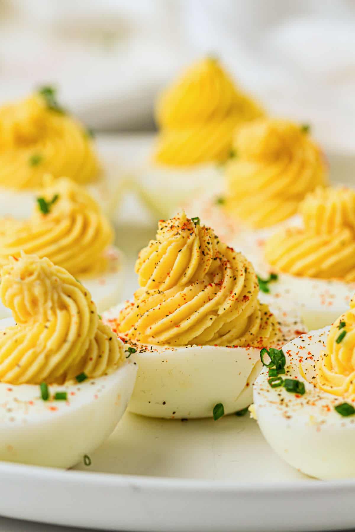 Devuled eggs are arranged on a white plate, with creamy yellow filling piped into swirls, then garnished with chopped chives and a sprinkle of paprika.