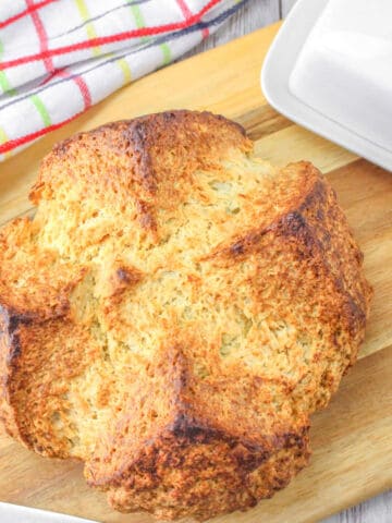 Round loaf of golden brown soda bread on a wooden cutting board, next to a white butter dish and a colorful checked kitchen towel.