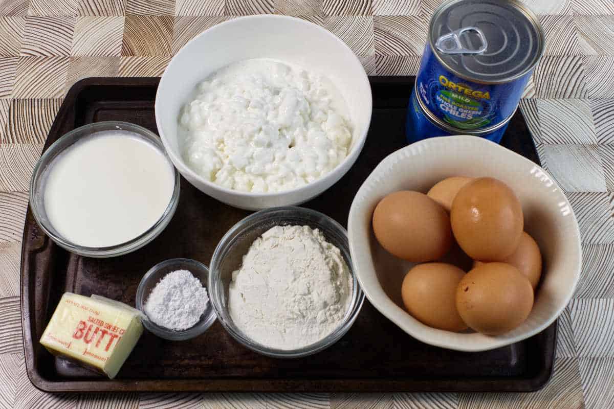 A baking tray holds bowls and containers with eggs, cottage cheese, flour, baking powder, milk, a stick of butter, and a can of chopped green chiles, arranged on a patterned surface.