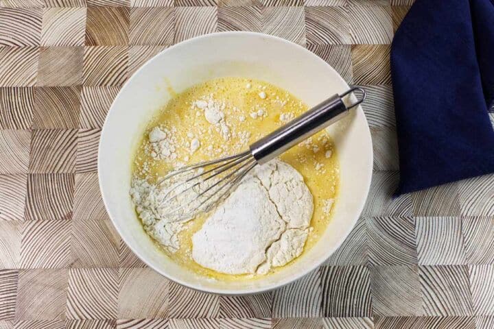 A white mixing bowl containing a yellow batter and a pile of flour, with a metal whisk resting inside, sits on a patterned wooden surface next to a dark blue cloth.