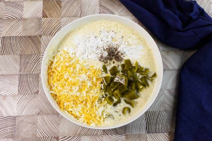 A white bowl containing shredded cheese, chopped green chilies, black pepper, and a creamy mixture, placed on a patterned wooden surface next to a dark blue cloth.