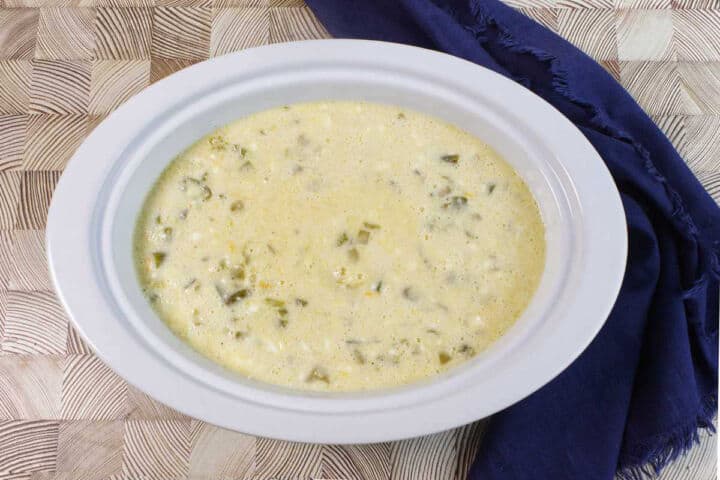 A white bowl filled with creamy, chunky soup sits on a patterned surface next to a dark blue cloth napkin. The soup contains visible green pieces, possibly vegetables or chiles.