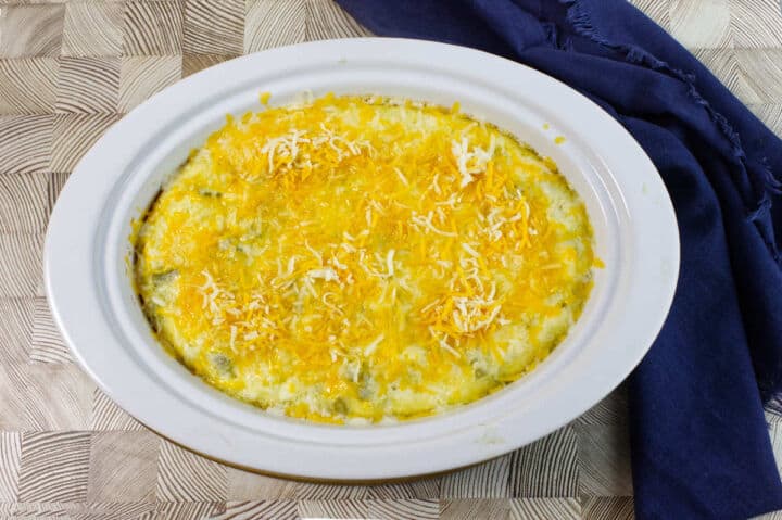 A white oval baking dish filled with a casserole topped with melted yellow and white shredded cheese, placed on a patterned wooden surface next to a dark blue cloth napkin.