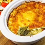 A cheesy casserole with a golden-brown top sits in a white slow cooker, with a serving spoon and a portion missing, revealing the creamy inside.
