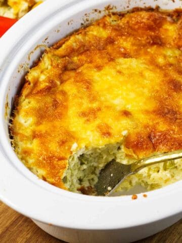 A cheesy casserole with a golden-brown top sits in a white slow cooker, with a serving spoon and a portion missing, revealing the creamy inside.