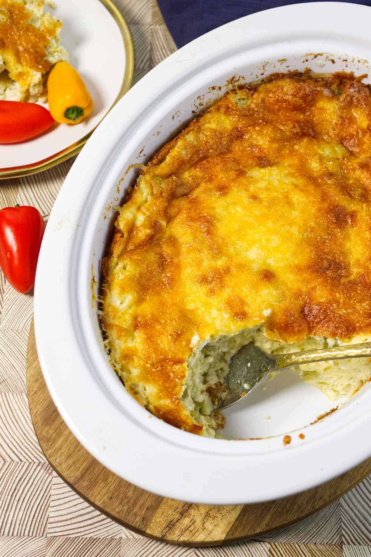 A baked casserole with a golden, cheesy crust in a white dish; a serving has been scooped out, revealing the creamy inside. A spoon rests in the dish, and colorful peppers are on a plate nearby.
