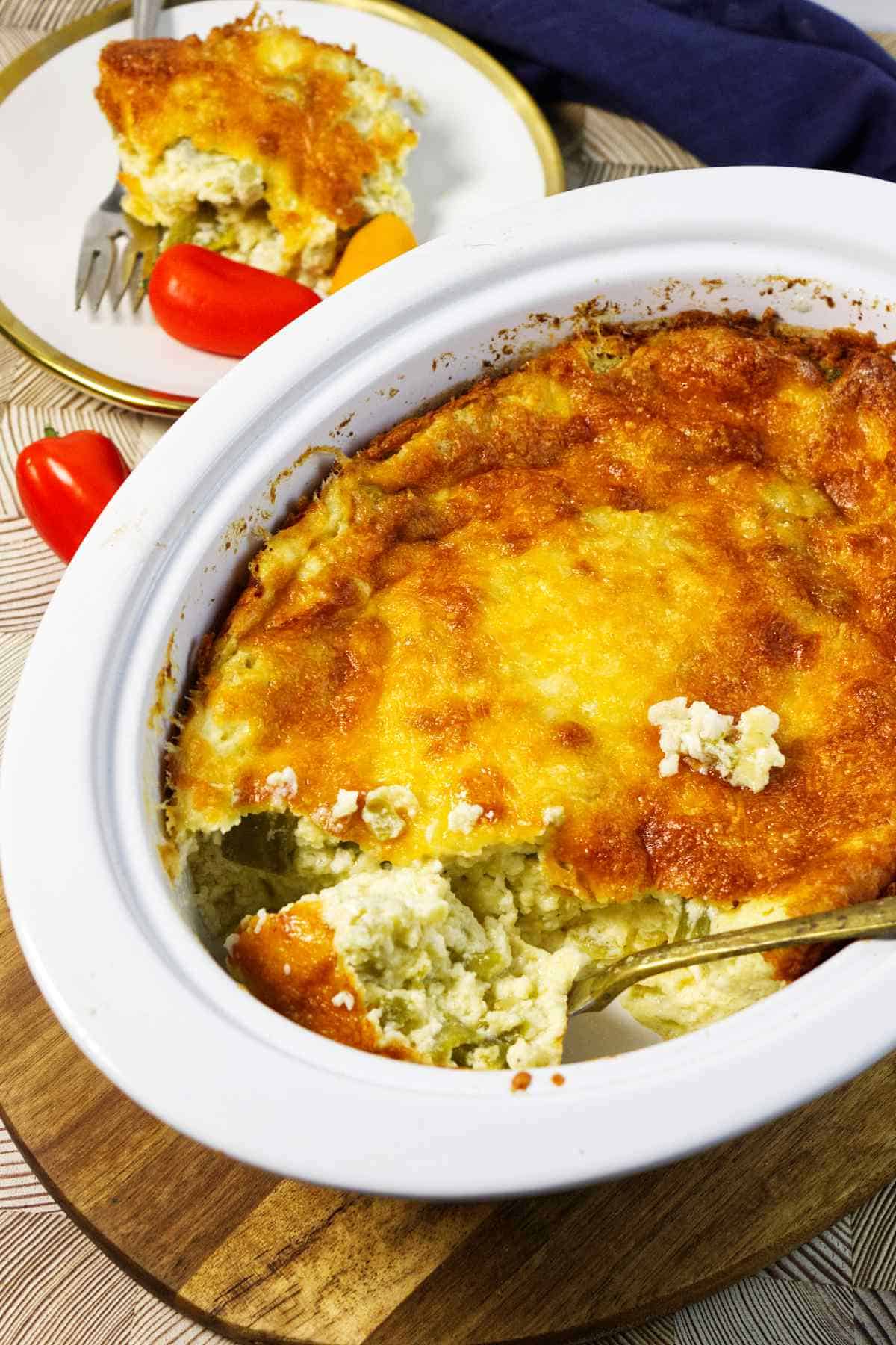 A baked cheesy casserole in a white dish with a golden brown top, a serving scooped out onto a plate in the background. Red mini bell peppers and a fork are next to the plate.