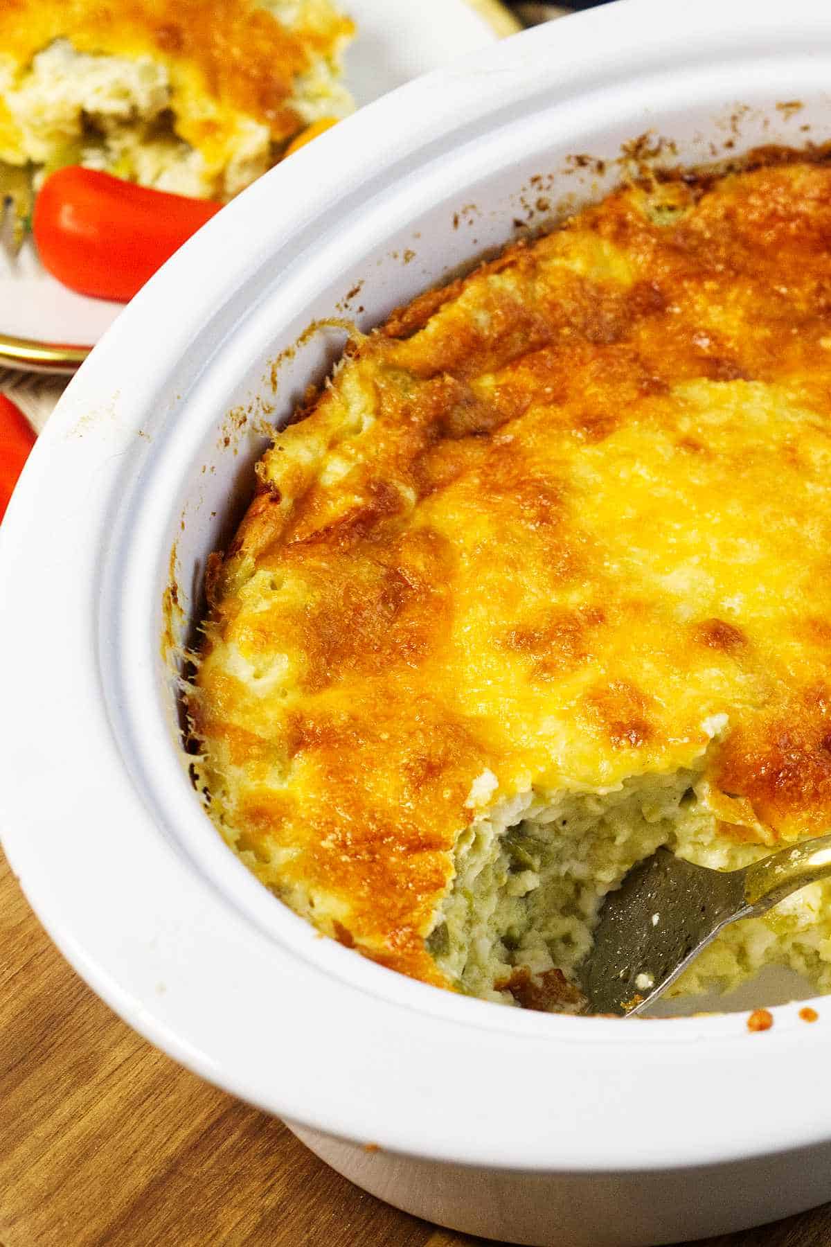 A baked casserole with a golden, cheesy crust sits in a white dish. A portion has been served, revealing a creamy interior. A spoon rests in the dish, and a plate with a serving and cherry tomatoes is visible in the background.
