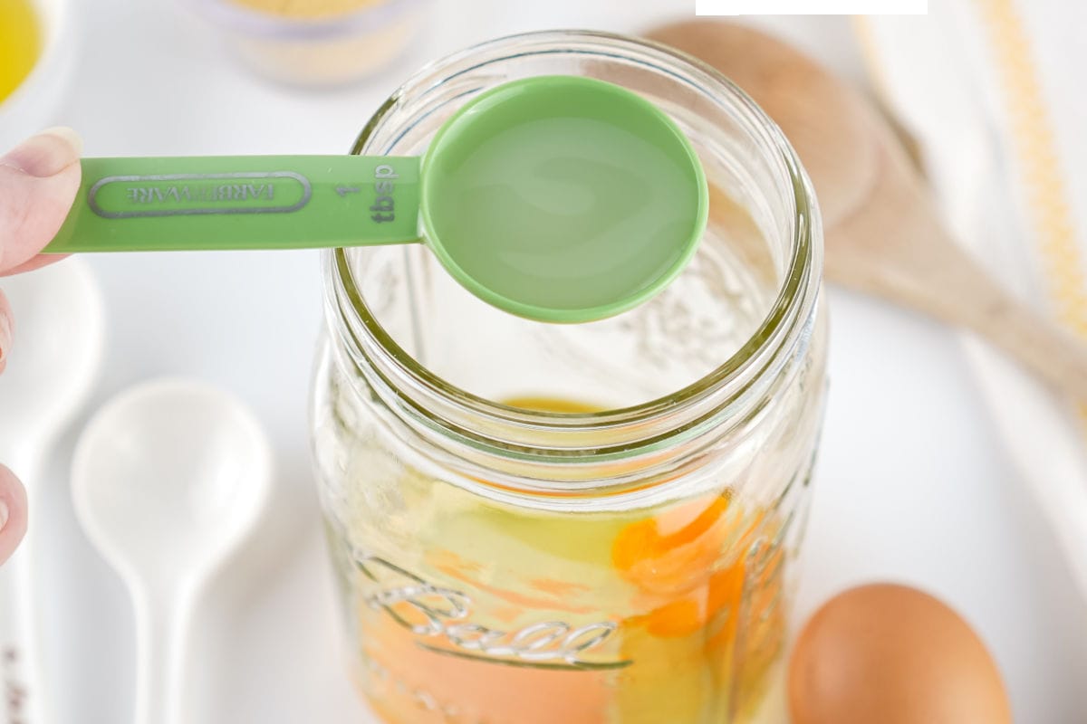 A green measuring spoon with liquid, likely for homemade mayonnaise, is held over a glass jar containing eggs and other ingredients. White measuring spoons, a wooden spoon, and an egg are nearby on a white surface.