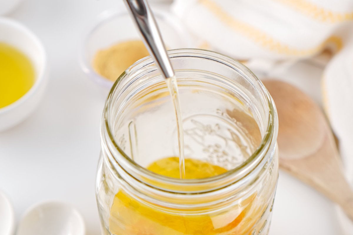 A close-up of a metal spoon lifting honey from a glass jar, surrounded by small bowls with yellow liquid, homemade mayonnaise, and a wooden spoon in the background on a white surface.