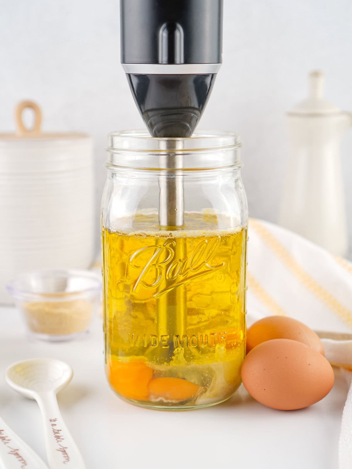 A black immersion blender is placed in a glass mason jar containing oil and eggs, ready to whip up homemade mayonnaise. Two brown eggs, a white measuring spoon, a small bowl of seasoning, and a striped towel are nearby on a white surface.