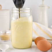A glass mason jar filled with creamy yellow homemade mayonnaise is being blended with an immersion blender. Nearby are two brown eggs, a white spoon, and a striped kitchen towel on the countertop.