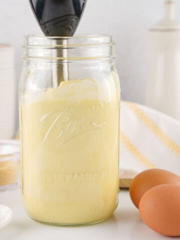 A glass mason jar filled with creamy yellow homemade mayonnaise is being blended with an immersion blender. Nearby are two brown eggs, a white spoon, and a striped kitchen towel on the countertop.
