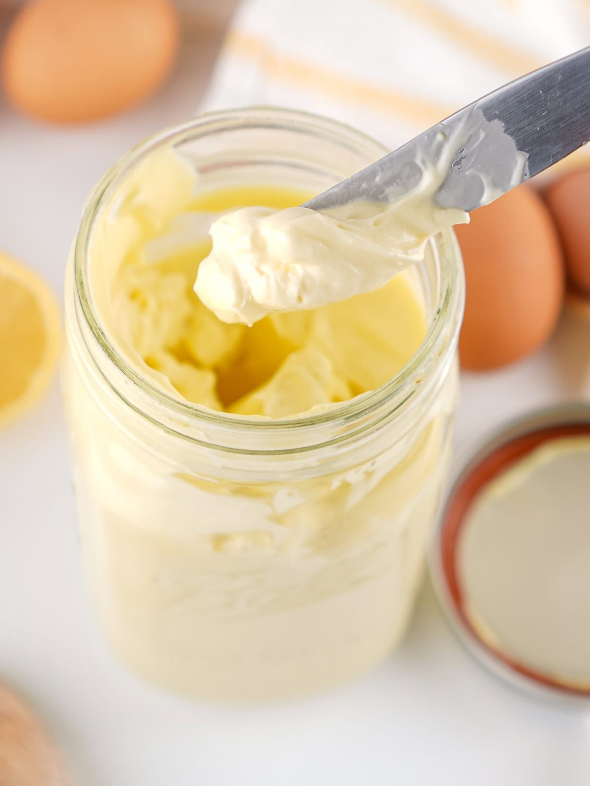 An open glass jar filled with creamy homemade mayonnaise, with a knife scooping some out. Fresh eggs, a lemon, and the jar lid are visible in the background.