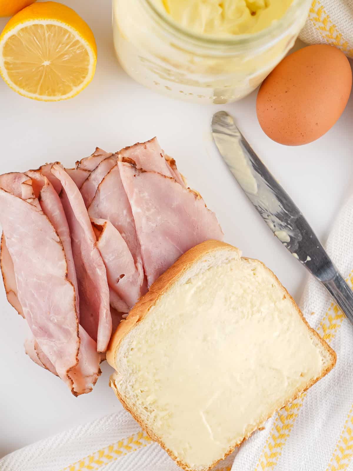 Slices of ham on a piece of bread, another bread slice spread with homemade mayonnaise, a knife, a whole egg, a halved lemon, and a jar of mayonnaise are arranged on a white surface with a yellow-striped cloth.
