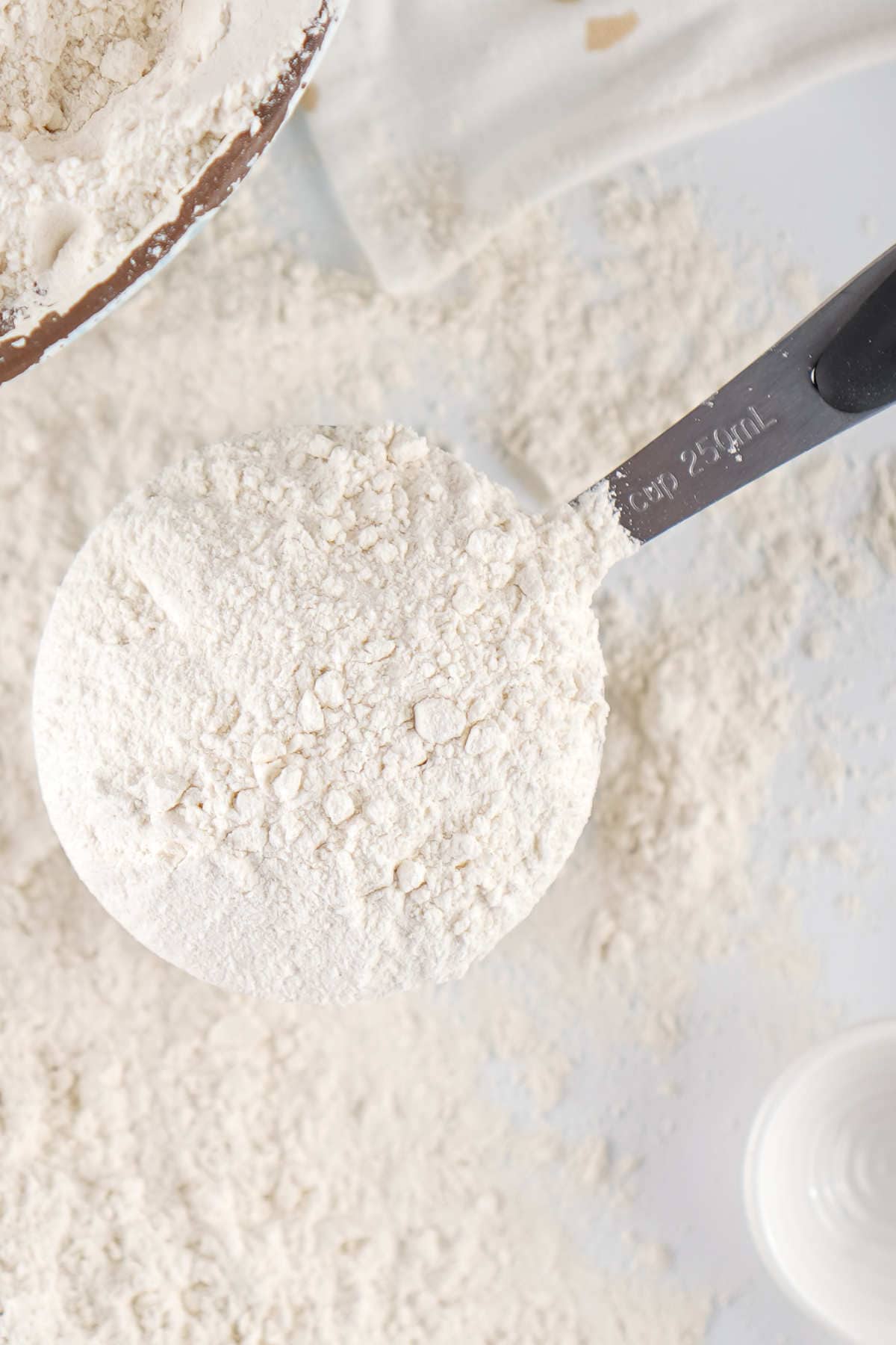 A metal measuring cup filled with flour rests on a white surface scattered with loose flour. A mixing bowl with more flour is partially visible in the top left corner.