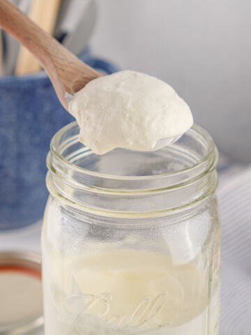 A wooden spoon holds a dollop of thick, creamy yogurt above an open glass mason jar, with a blue container and striped cloth blurred in the background.