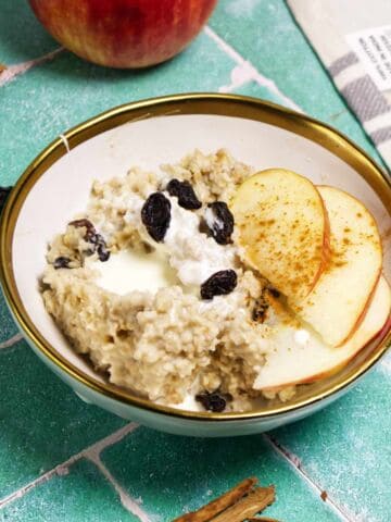 A bowl of oatmeal topped with apple slices, raisins, a drizzle of cream, and a sprinkle of cinnamon, placed on a green tiled surface with a whole apple and cinnamon sticks nearby.