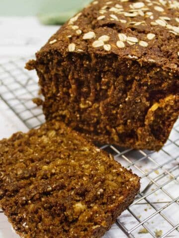 A loaf of brown bread topped with oats sits on a wire cooling rack. One slice is cut and lies in front of the loaf, showing the moist texture inside. The background is a light wooden surface.