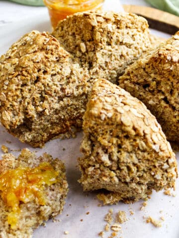 A rustic loaf of Irish wheaten bread is cut into wedges on parchment paper. One piece has orange marmalade spread on it, and a jar of marmalade sits in the background.