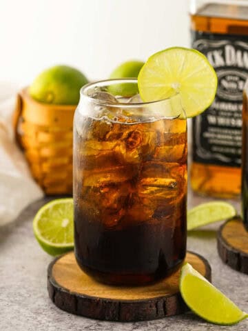 A glass of dark soda with ice and a lime wheel garnish, sitting on a wooden coaster. A bottle of Jack Daniel's whiskey and a basket of limes are in the background. Lime wedges are on the surface nearby.