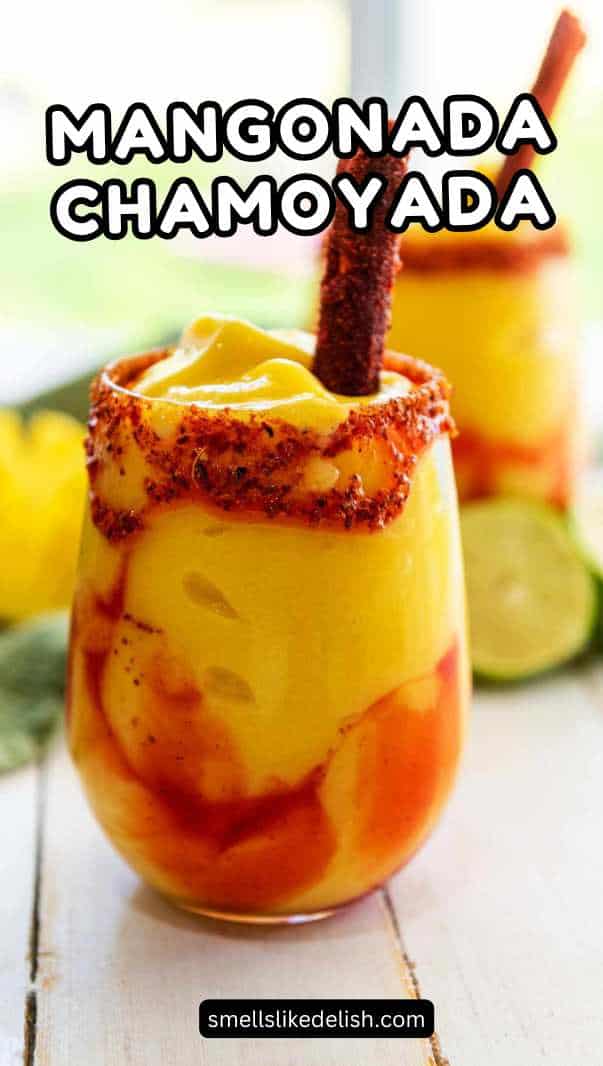 A glass of bright yellow Mangonada smoothie with red chamoy sauce swirls, topped with a tamarind candy stick and chili-lime seasoning on the rim, with another chamoyada, lime slices, and a green napkin in the background.