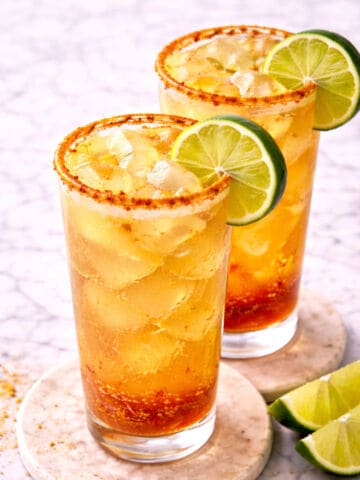 Two tall glasses filled with an amber-colored iced drink, garnished with lime slices and a chili powder rim. The glasses are placed on coasters, with extra lime wedges nearby, on a light marble surface.