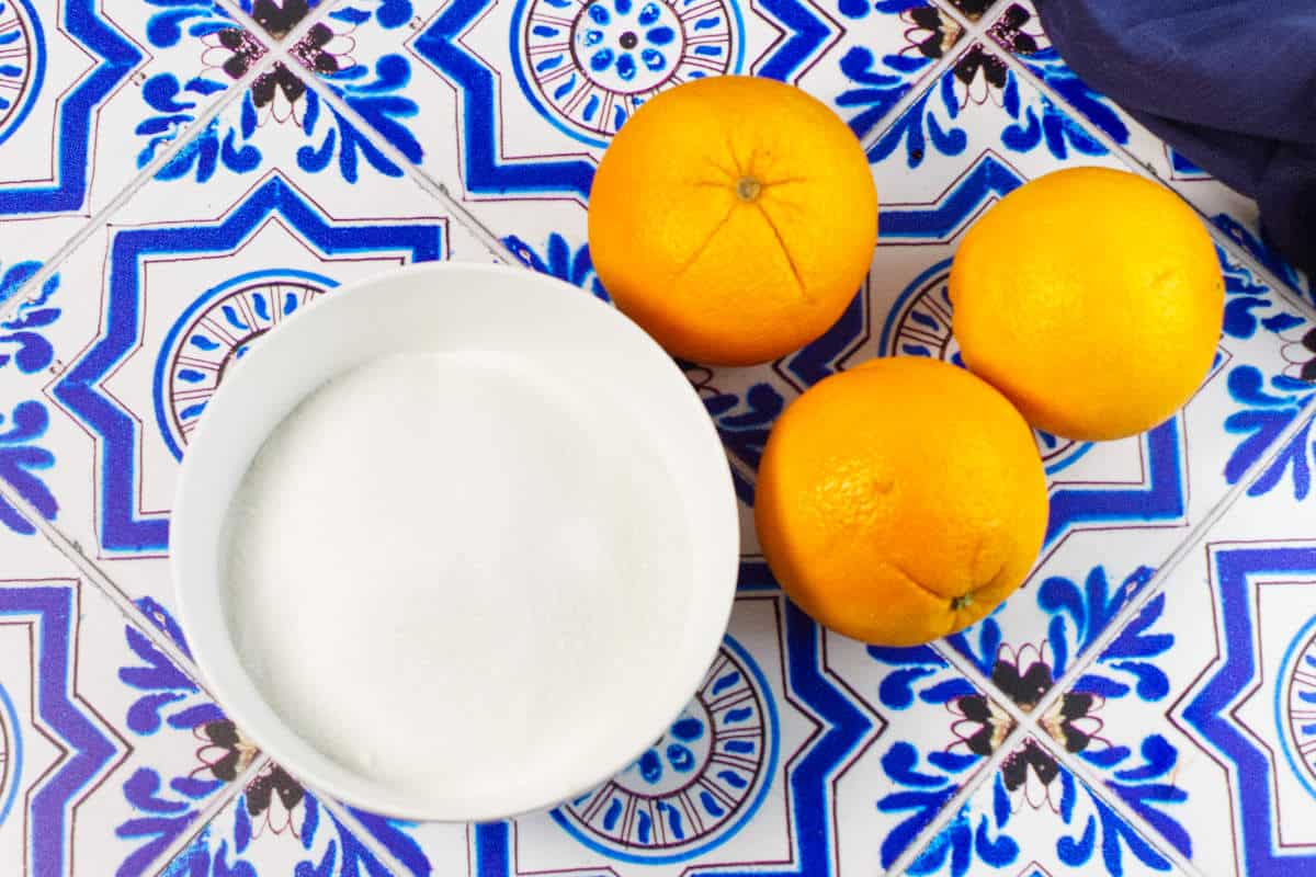A white bowl filled with sugar sits next to three whole oranges on a blue and white patterned tile surface, perfect ingredients for homemade orange marmalade.