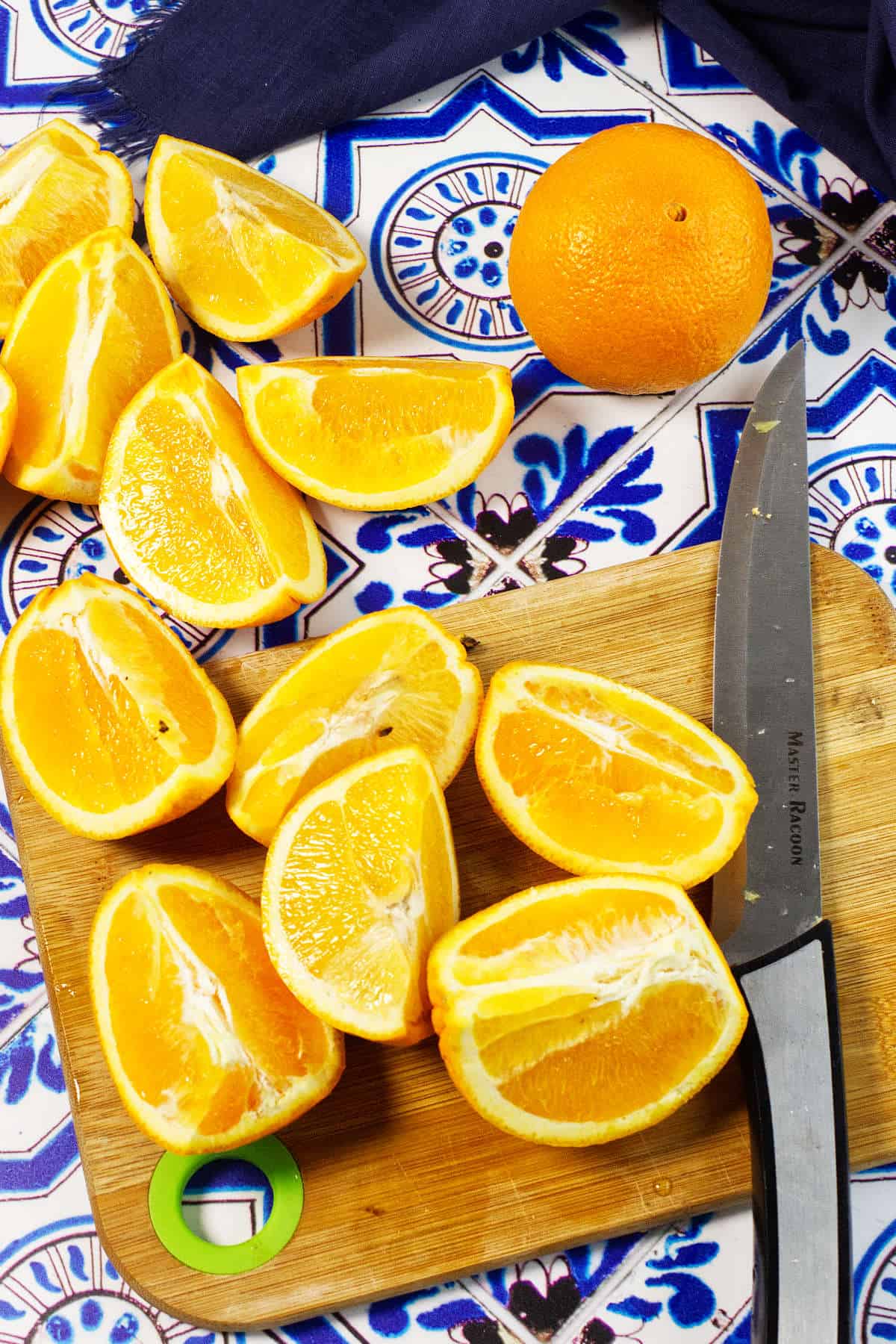 Sliced oranges on a wooden cutting board with a knife, perfect for making orange marmalade, alongside extra wedges and a whole orange on a patterned blue and white tile surface.
