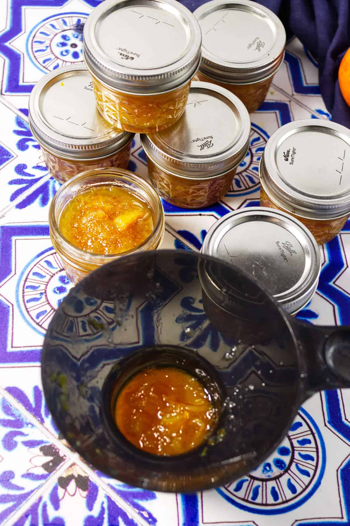 Small mason jars filled with orange marmalade are stacked on a blue and white patterned tile surface. In the foreground, an open jar and a black ladle showcase the vibrant, homemade orange marmalade.