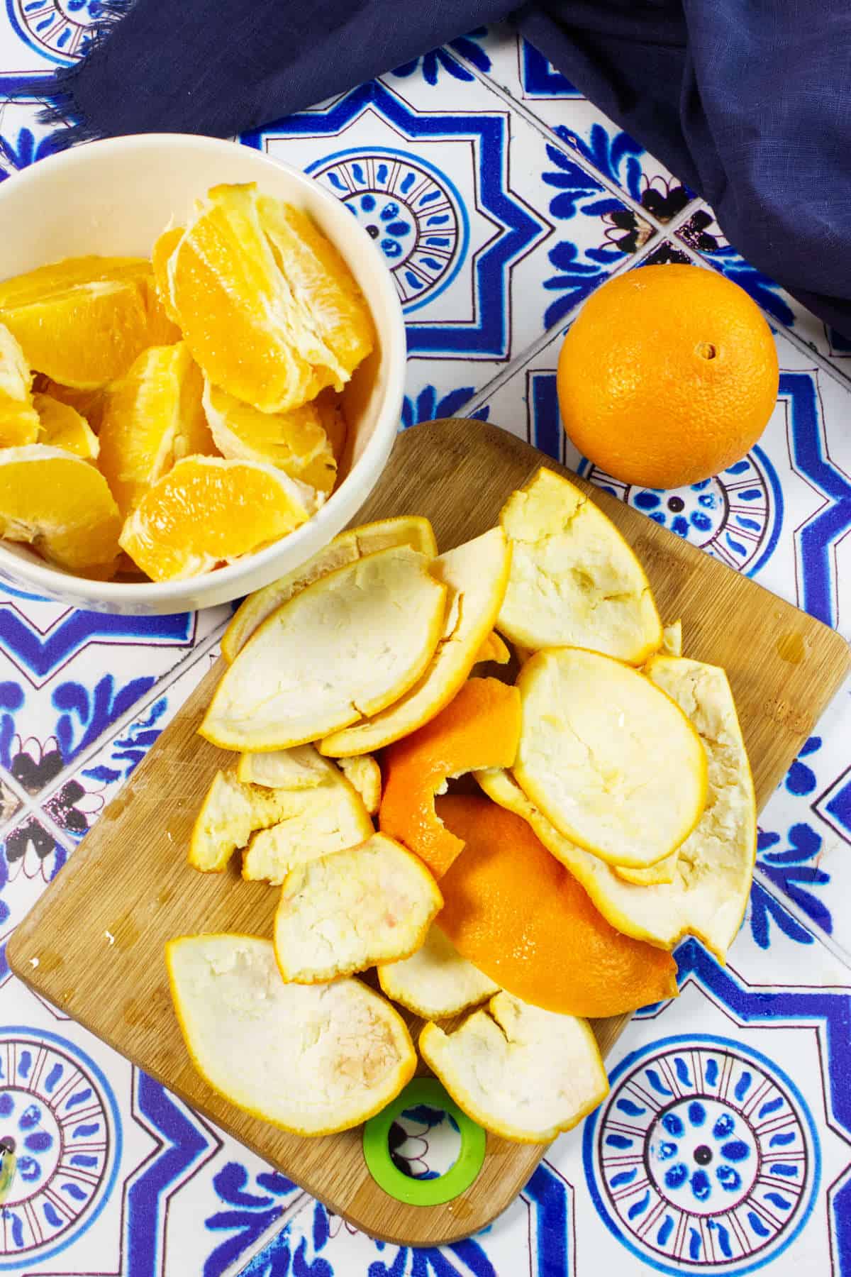 A bowl of peeled orange segments, orange peels on a wooden board, and a whole orange rest on a blue and white patterned surface, evoking the freshness perfect for making homemade orange marmalade.