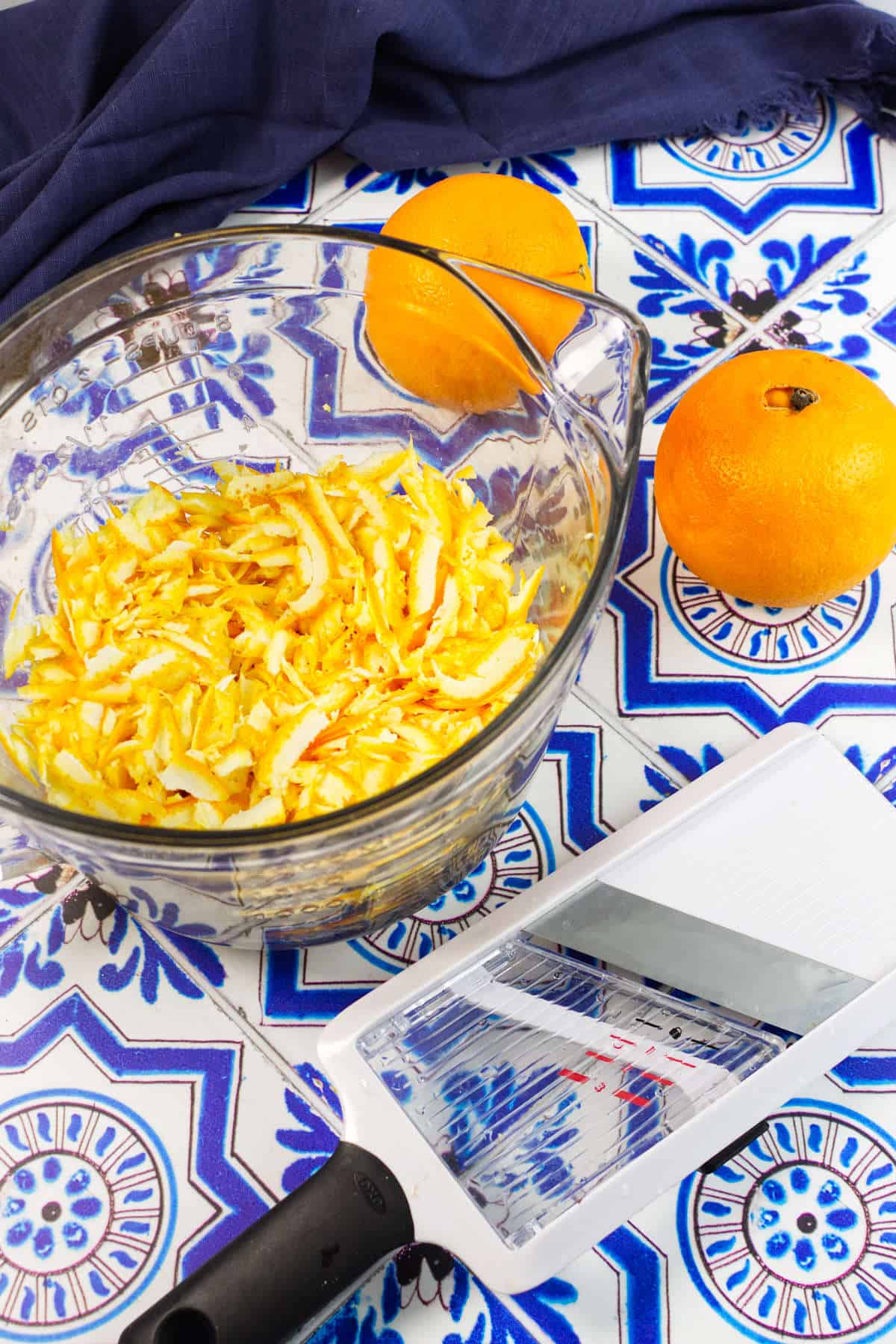 A glass bowl filled with shredded orange and white fruit, perfect for making orange marmalade, sits on a blue and white patterned surface. Beside it are two whole oranges and a mandoline slicer with juice and zest on the blade.