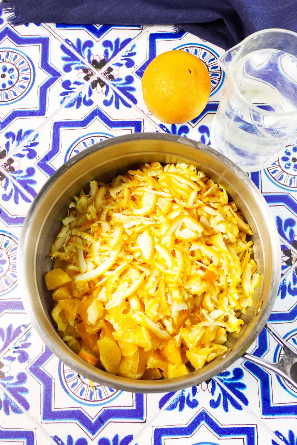 A metal bowl filled with chopped oranges and peels, perfect for making orange marmalade, sits on a blue and white patterned surface. Nearby are a whole orange and a clear pitcher of water.