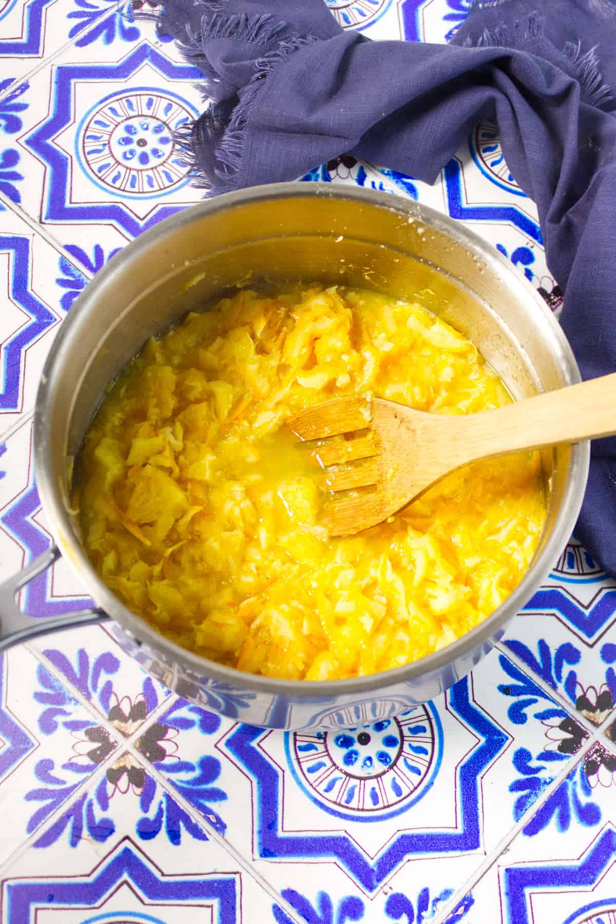 A pot of bright yellow mango jam, reminiscent of orange marmalade, is being stirred with a wooden spatula on a colorful blue and white patterned tile surface, with a dark blue cloth nearby.