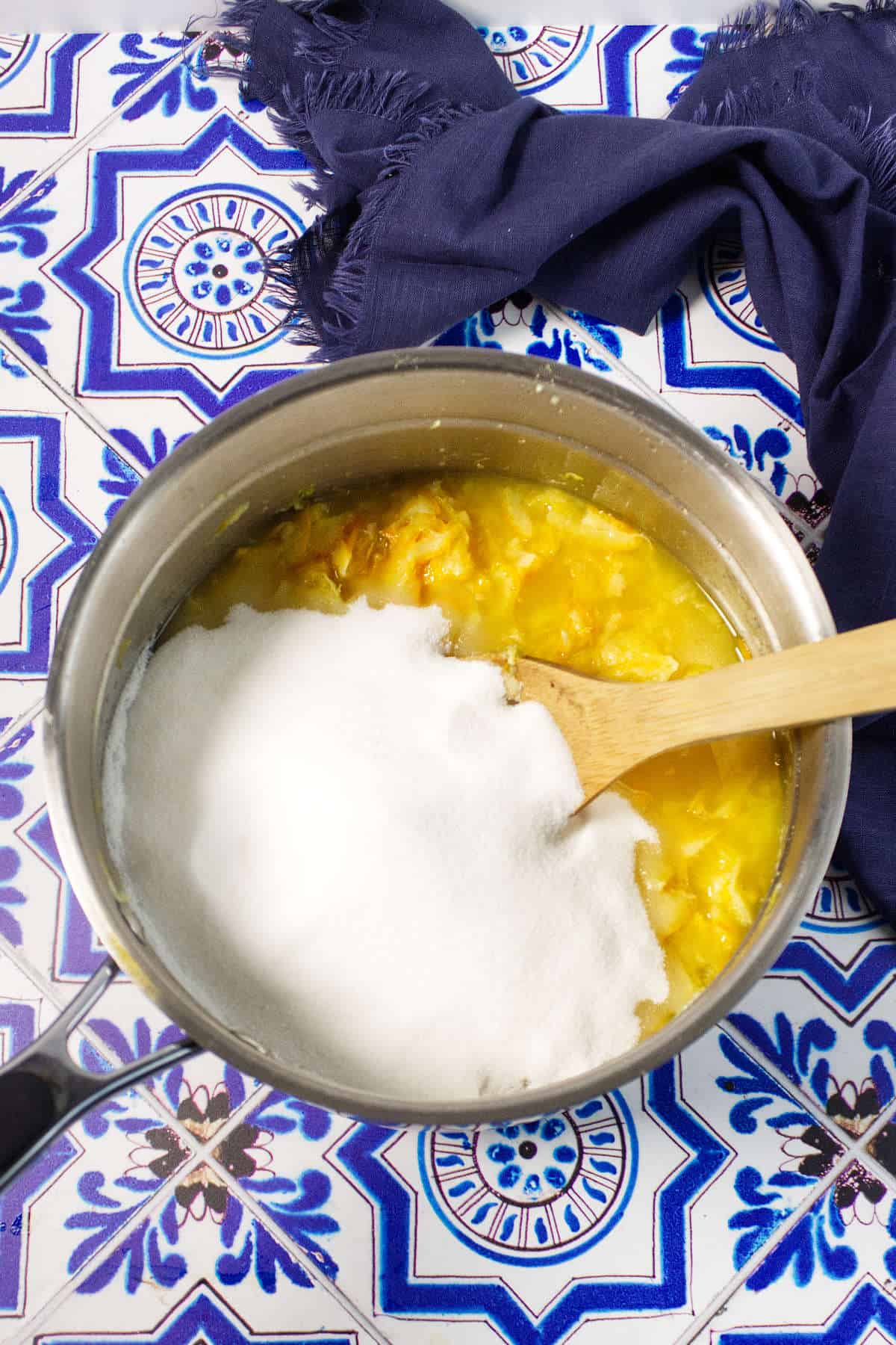 A saucepan with an orange marmalade mixture and a pile of white sugar on top, being stirred with a wooden spoon, sits on a blue and white patterned tile surface with a dark blue cloth nearby.