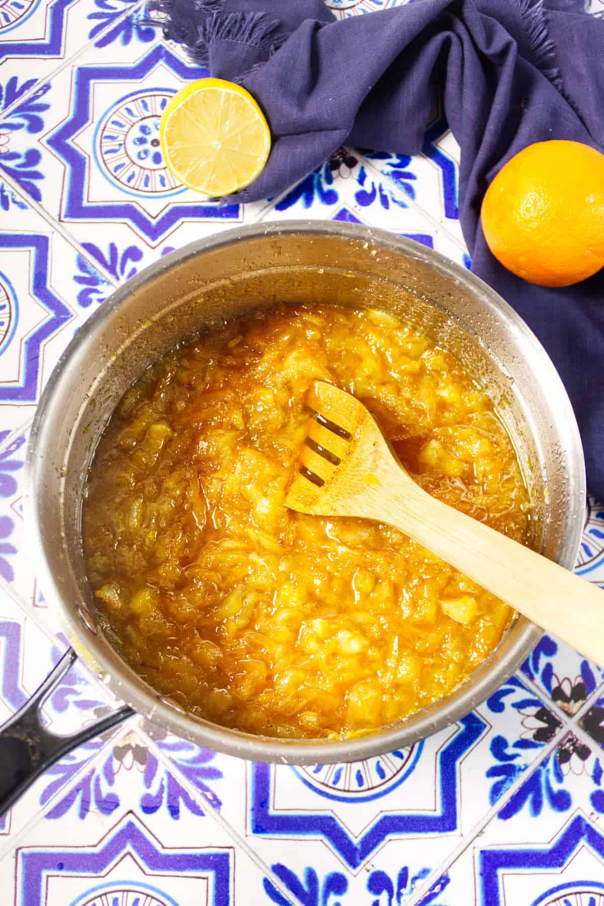 A pot of orange marmalade being stirred with a wooden spoon sits on a blue patterned tile surface, with a halved lemon, fresh orange, and dark blue cloth nearby.