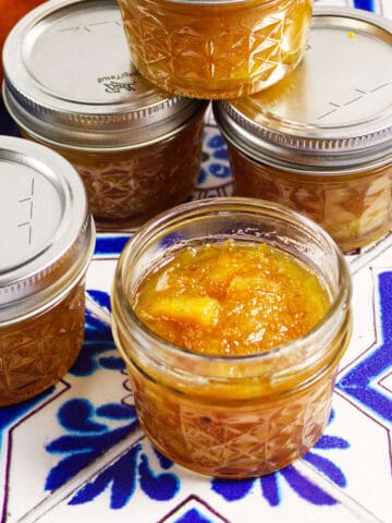 Five small glass jars filled with zesty orange marmalade sit on a blue and white patterned tile surface; one jar is open, revealing the chunky spread inside.