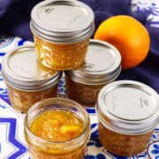 Five small glass jars filled with orange marmalade are arranged on a blue and white patterned surface, with one jar open in front. A whole orange sits in the background, and a dark blue cloth is partially visible behind the jars.