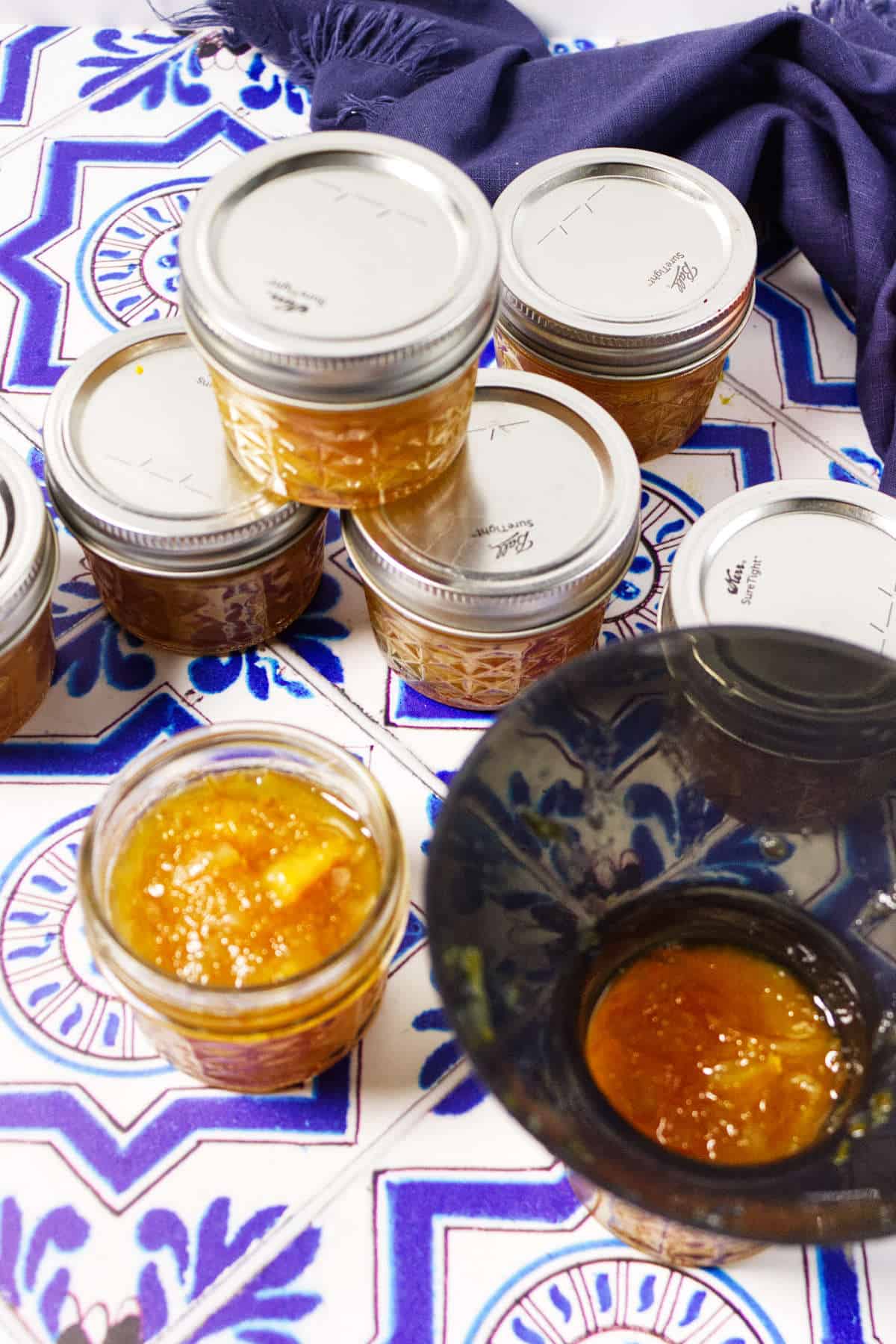 Small glass jars filled with vibrant orange marmalade are stacked on a blue and white patterned tablecloth, with a deep blue bowl partially visible in the foreground. A folded cloth napkin lies in the corner.